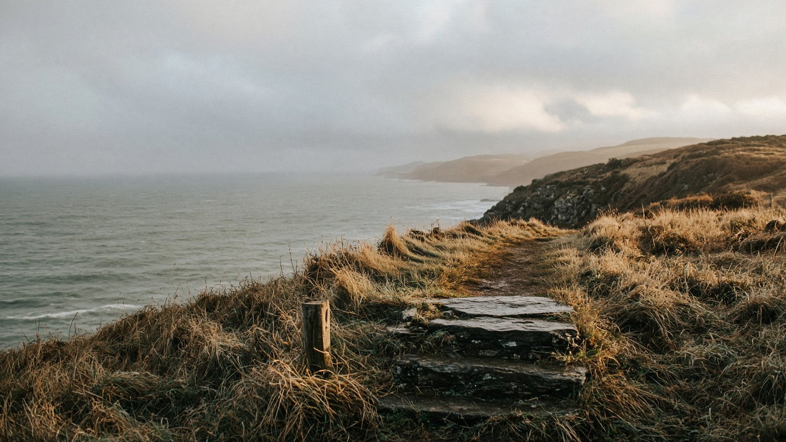 Coastal footpath above a grey-green sea, with a stone stile in the foreground