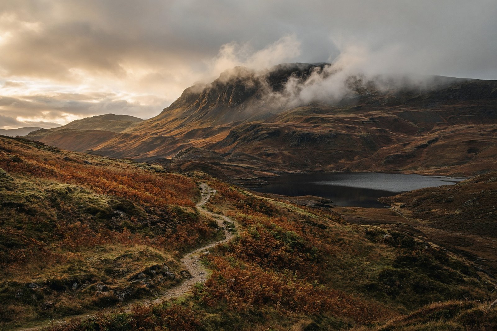 Fell path over bracken toward a dark tarn under breaking cloud